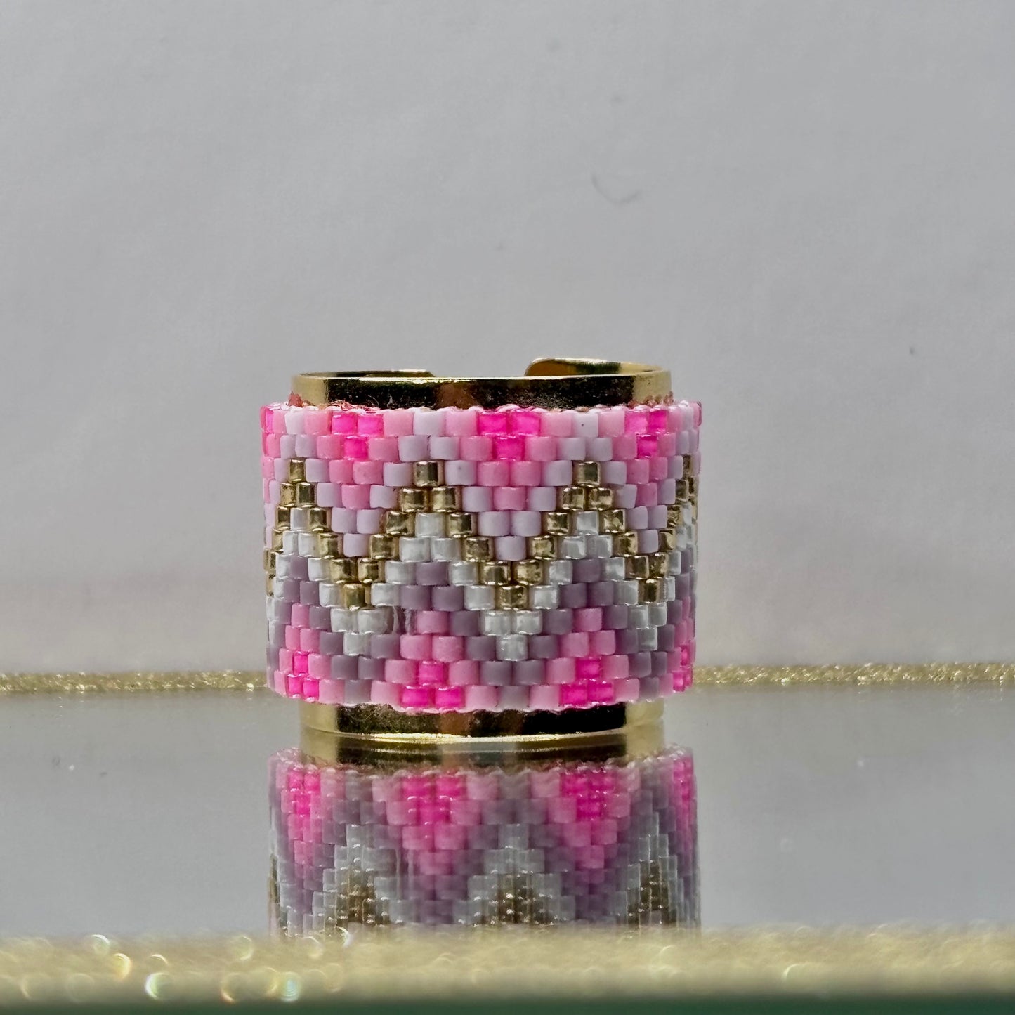 Beaded ring with pink, gold, and gray pattern on a reflective surface