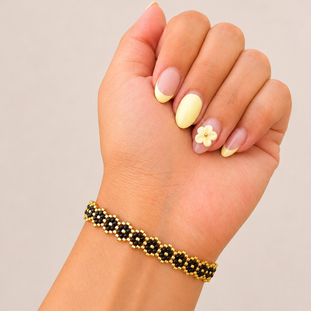 A close-up of a person's hand and wrist wearing a beaded bracelet. The bracelet features alternating black flower-shaped beads with gold centers and gold outer beads.