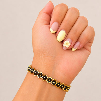 A close-up of a person's hand and wrist wearing a beaded bracelet. The bracelet features alternating black flower-shaped beads with gold centers and gold outer beads.