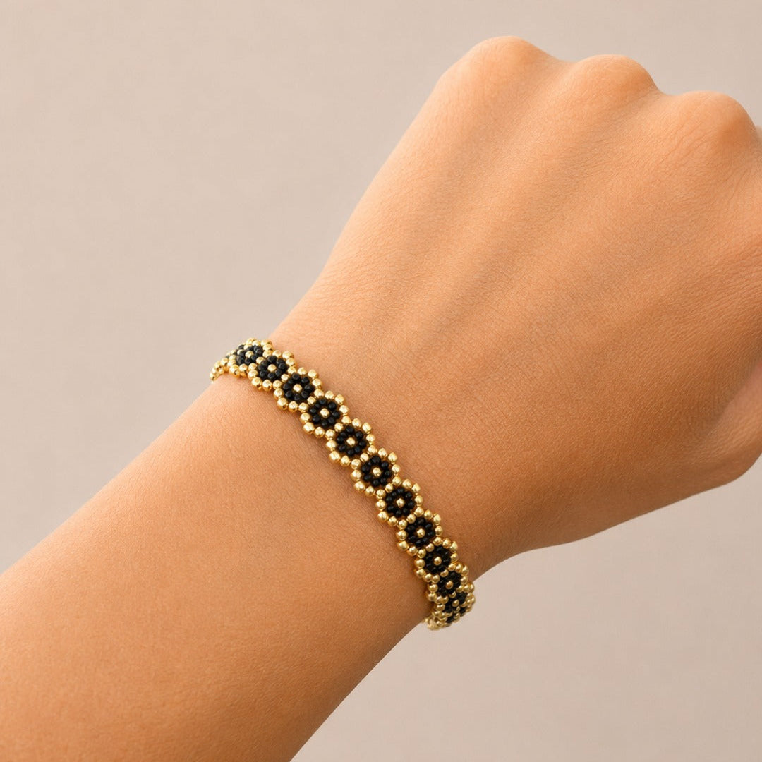 A gold beaded bracelet with black flower-shaped beads, shown worn on a person's wrist against a plain beige background.