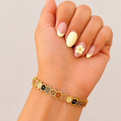 A close-up of a hand wearing a delicate beaded bracelet. The bracelet features a gold-tone base with small black, white, and orange beads arranged in a floral pattern.