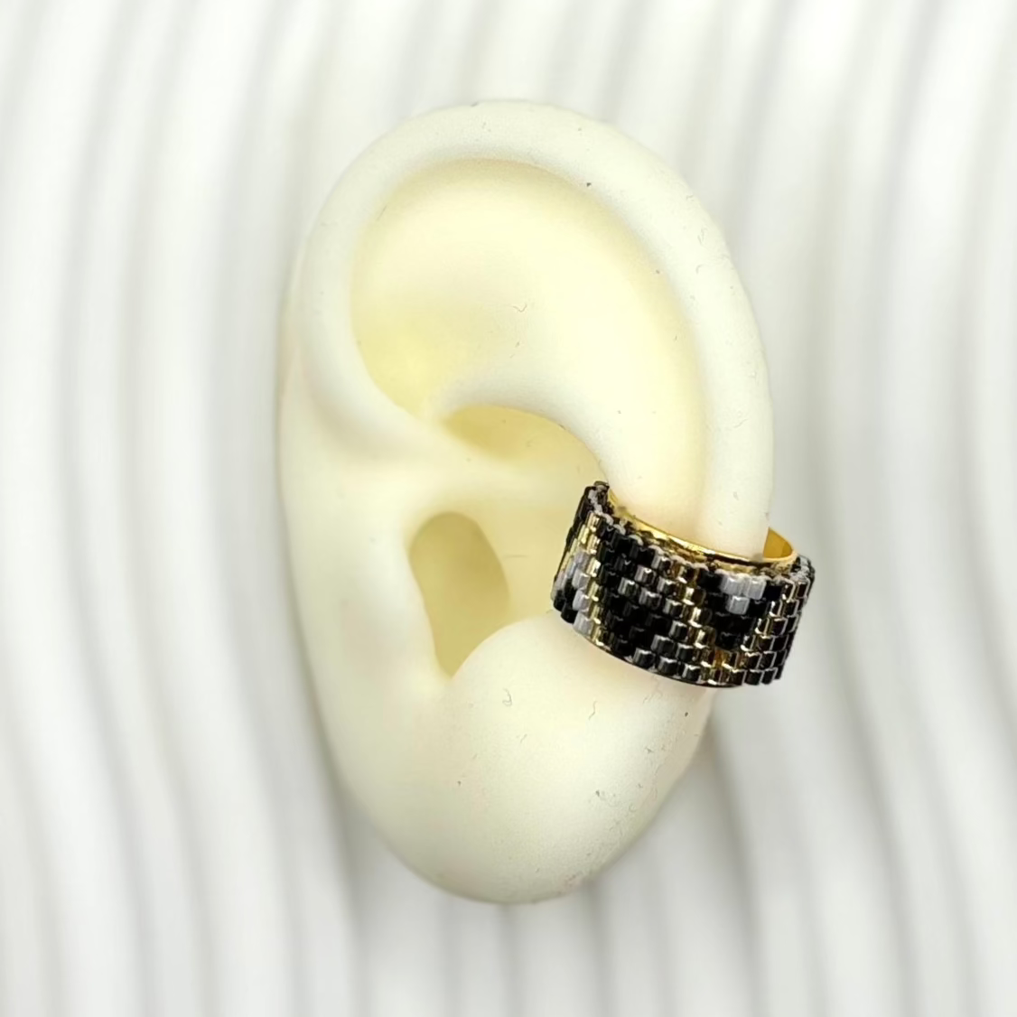 An ear cuff featuring black and gold Miyuki triangles, displayed on a white ear mold to show fit.