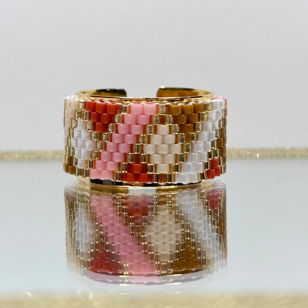 Beaded ring with pink, white, and gold pattern on a reflective surface