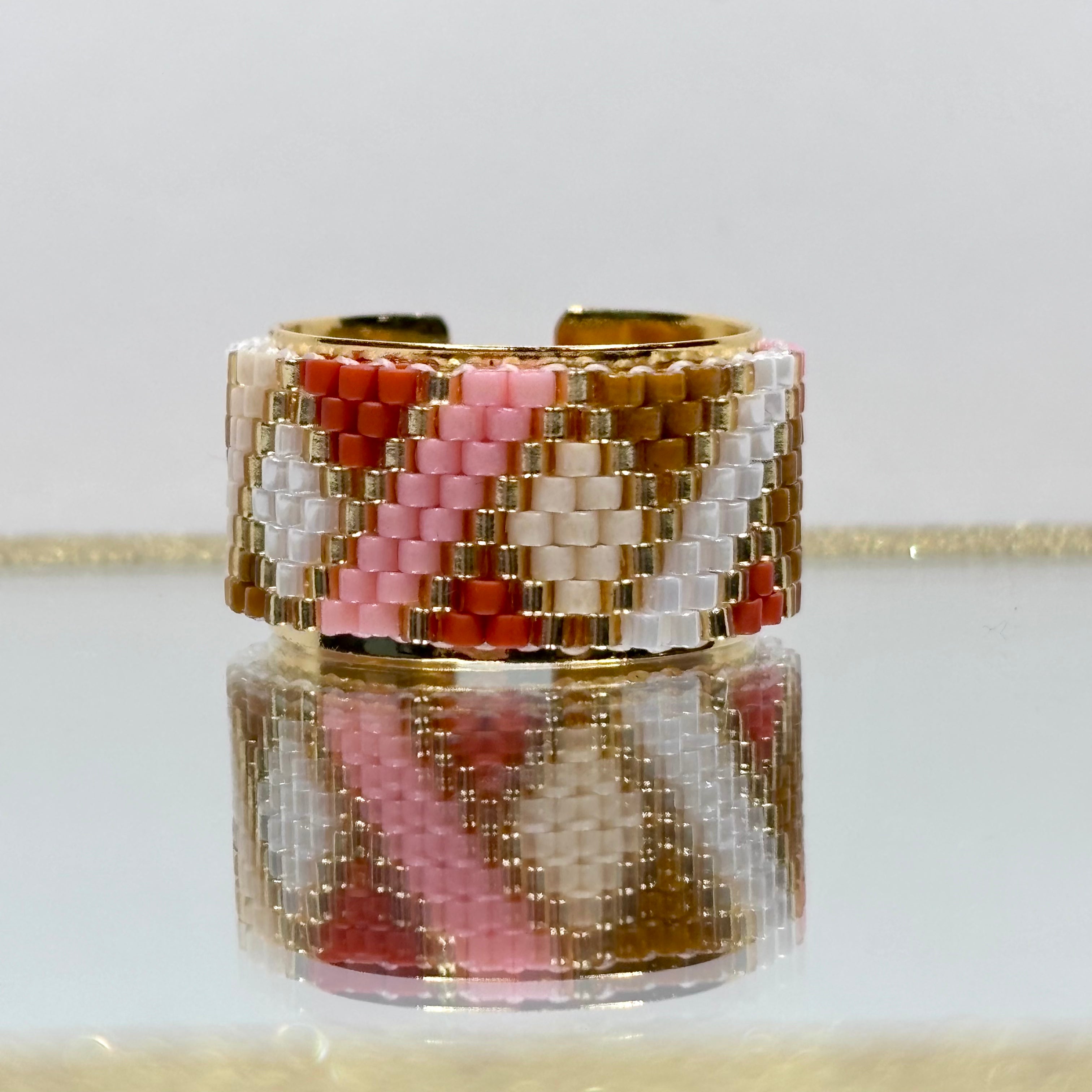 Beaded ring with pink, white, and gold pattern on a reflective surface