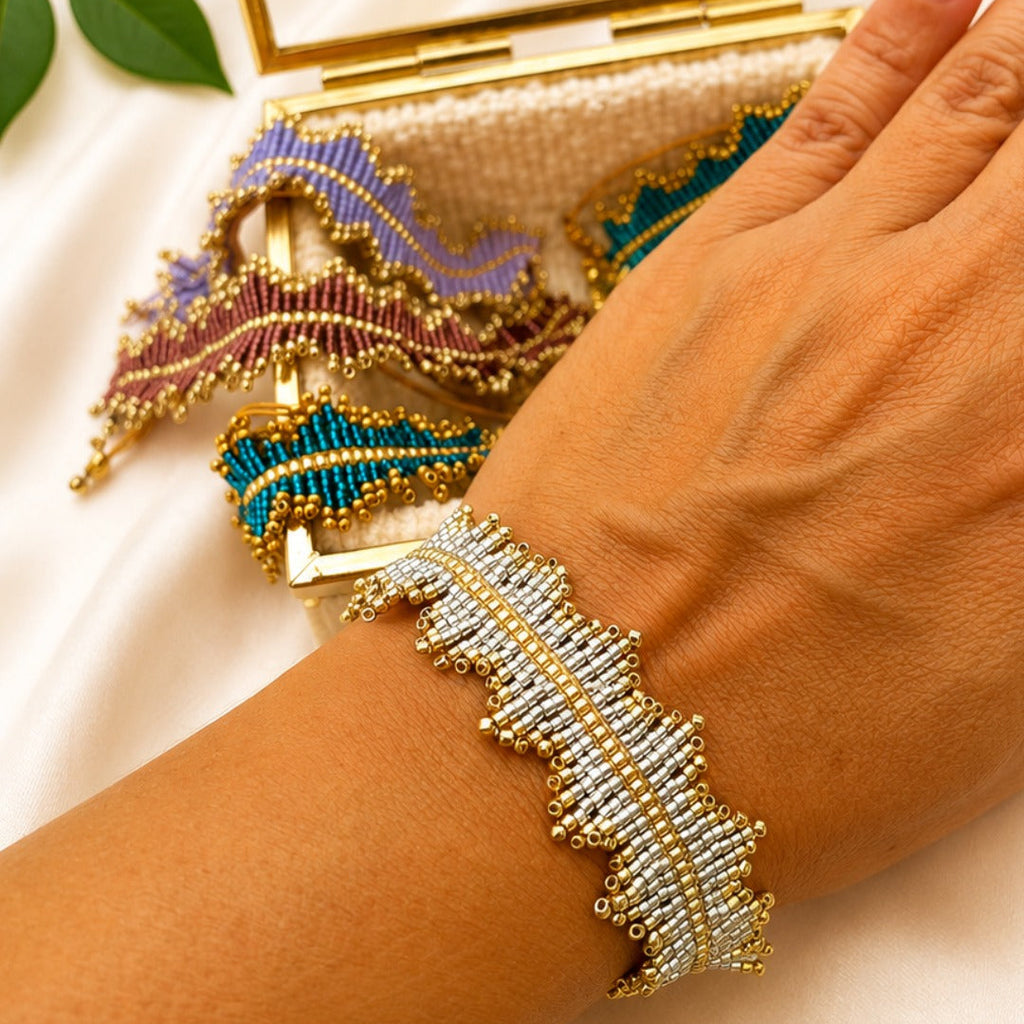 A close-up of a hand wearing a wide, beaded cuff bracelet with a gold-toned border and white beadwork. A matching multicolor beaded clutch purse with purple, teal, and gold details rests behind the hand on a white satin fabric background.