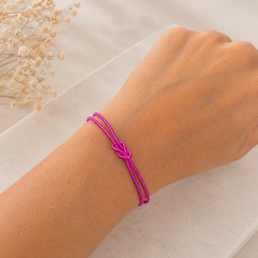 A close-up of a person's wrist wearing a bright magenta pink braided cord bracelet with a knotted detail, resting on a light beige stone surface with dried flowers in the background.