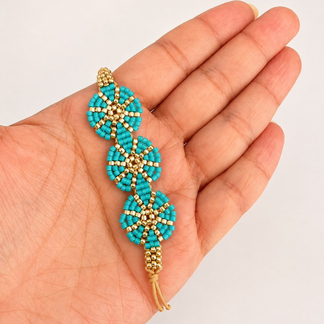 A handmade beaded bracelet with three turquoise and gold floral motifs, held in a person's hand against a plain light background.