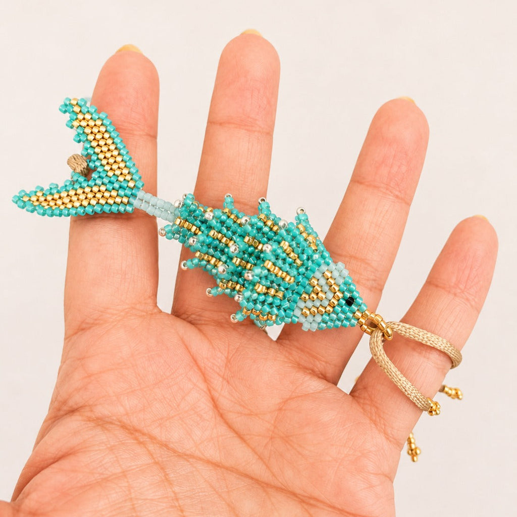 A turquoise and gold beaded fish-shaped ring held on a person's hand against a plain light background.