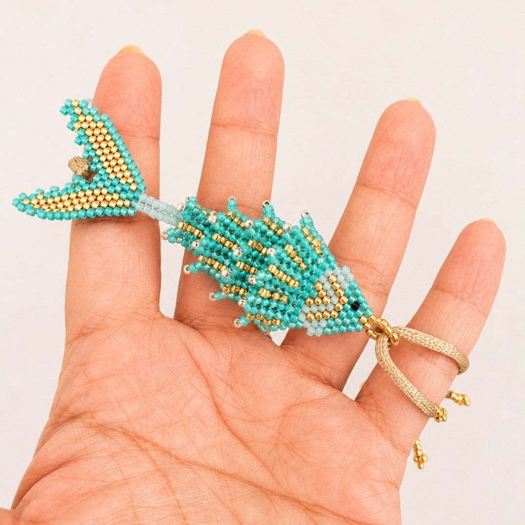 A turquoise and gold beaded fish-shaped ring held on a person's hand against a plain light background.