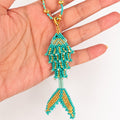 A turquoise and gold beaded fish-shaped pendant or charm displayed on a person's hand against a plain white background.