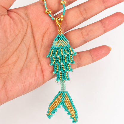 A turquoise and gold beaded fish-shaped pendant or charm displayed on a person's hand against a plain white background.