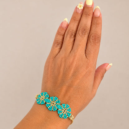 A close-up of a person's hand wearing a turquoise and gold beaded bracelet against a plain light gray background.