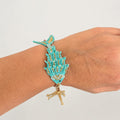 A turquoise and gold beaded bracelet designed to look like a coral reef with a dangling gold starfish, shown worn on a person's wrist against a plain light gray background.
