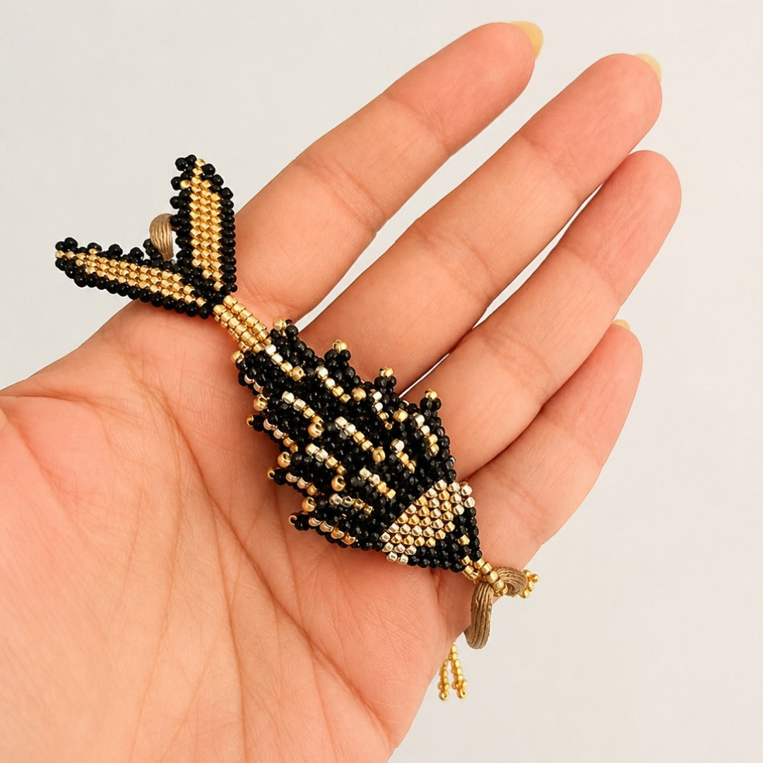 A hand holding a small beaded fish charm or brooch in black and gold seed beads against a plain light background.
