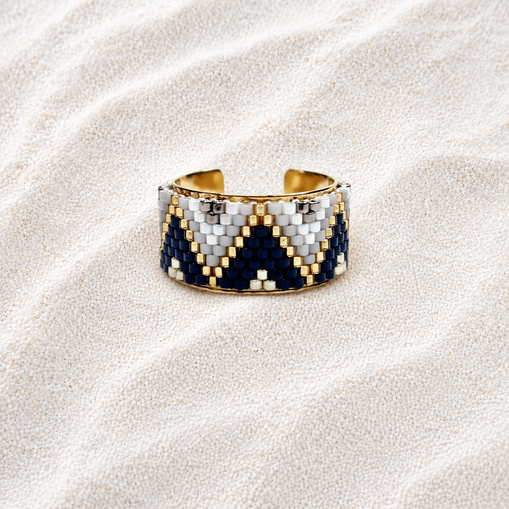 A wide cuff bracelet with a geometric beaded pattern in navy blue, white, and gold, displayed on a textured beige sand background.