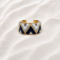 A wide cuff bracelet with a geometric beaded pattern in navy blue, white, and gold, displayed on a textured beige sand background.