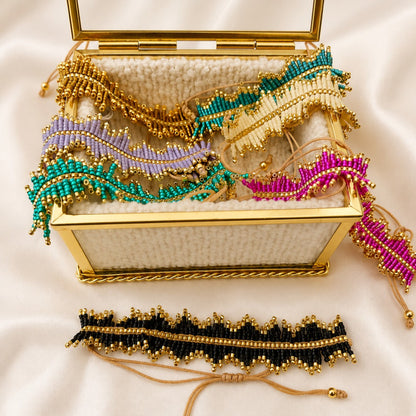 A gold-framed woven jewelry box filled with colorful beaded bracelets, with one black and gold beaded bracelet displayed in front on a cream satin background.