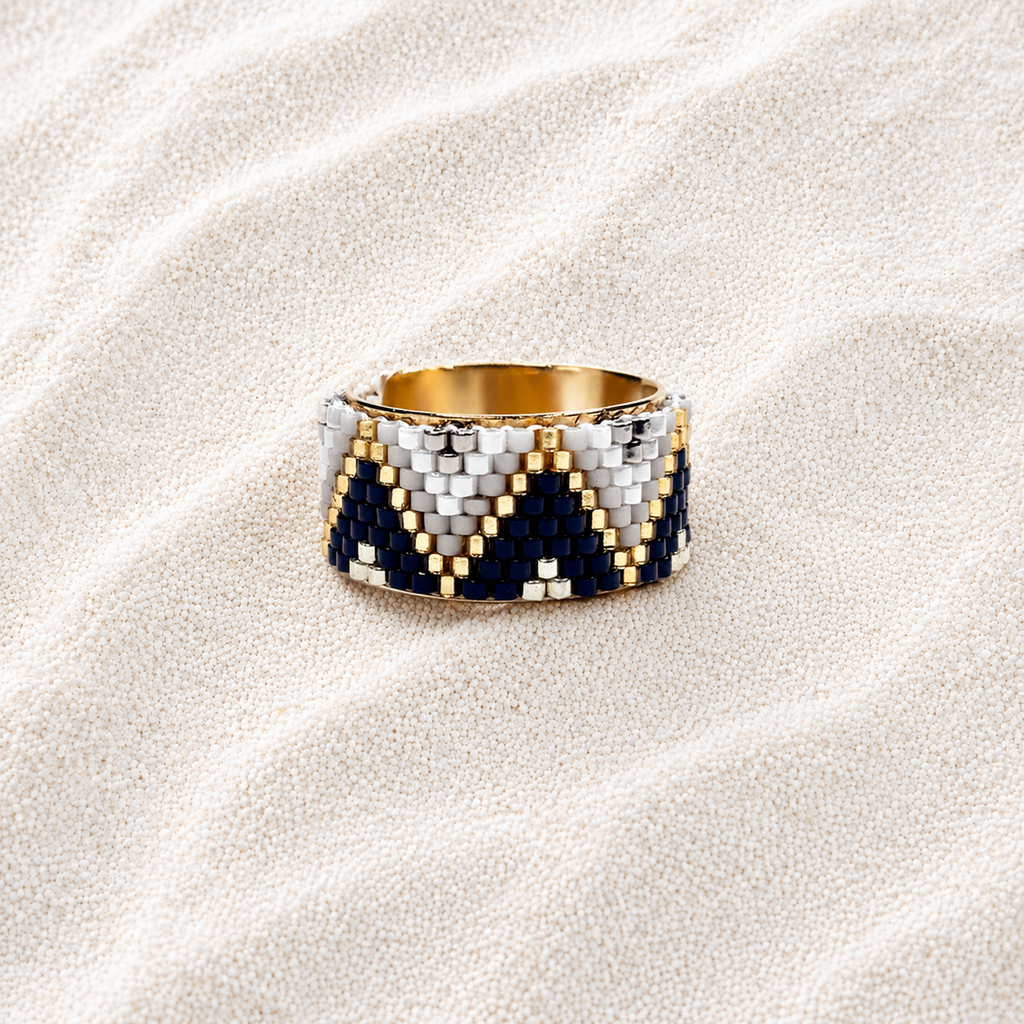 A wide beaded ring with a geometric chevron pattern in navy blue, white, and gold, displayed on a textured beige fabric background.