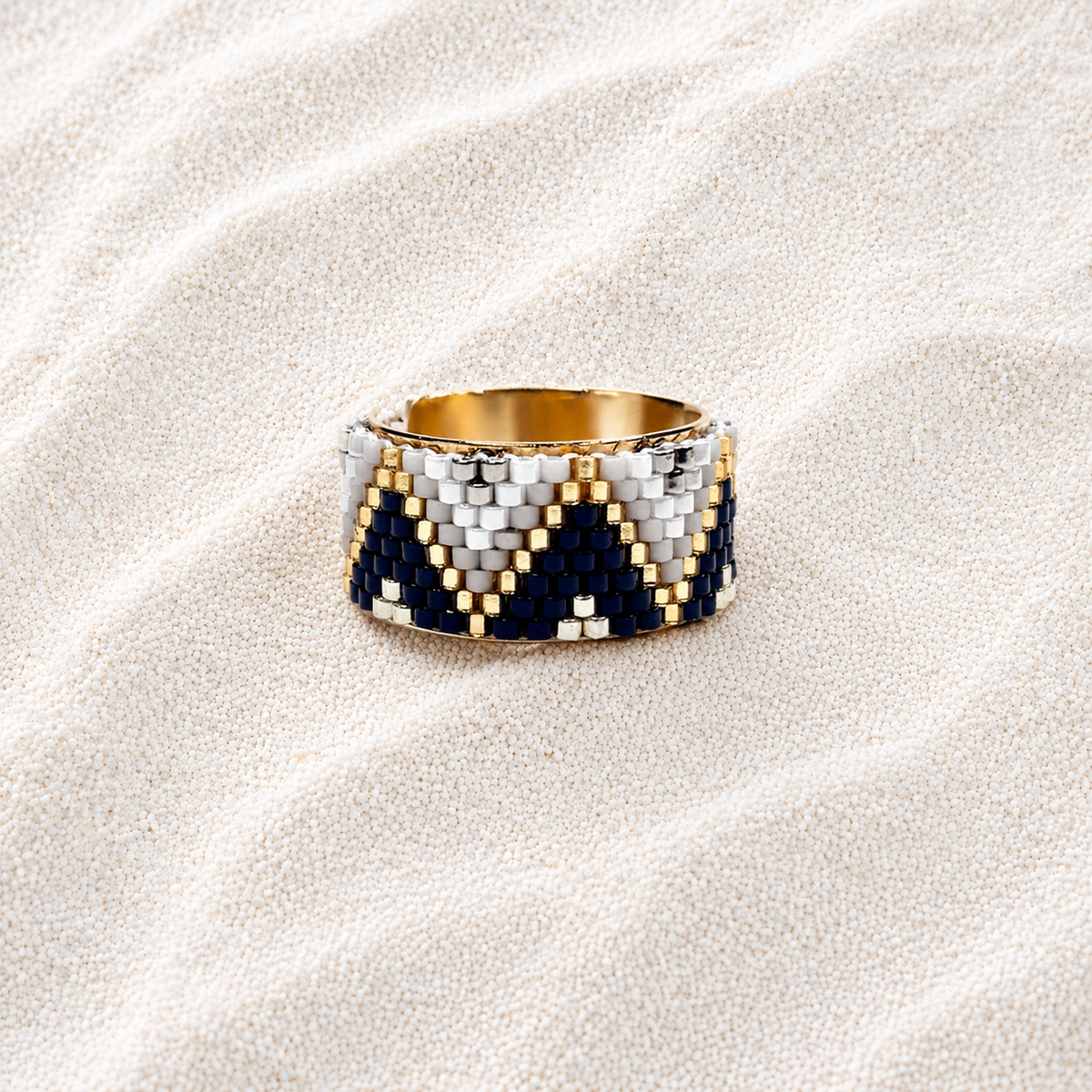 A wide beaded ring with a geometric chevron pattern in navy blue, white, and gold, displayed on a textured beige fabric background.