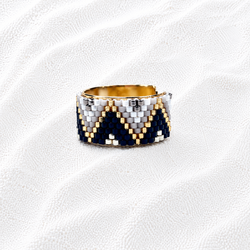 A wide band ring with a geometric chevron pattern made of navy blue, white, and gold seed beads, displayed on a textured white fabric background.