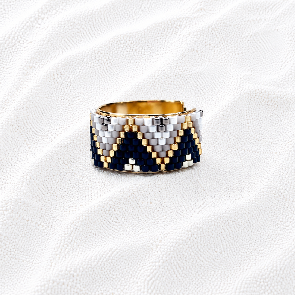 A wide band ring with a geometric chevron pattern made of navy blue, white, and gold seed beads, displayed on a textured white fabric background.