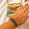A close-up of a hand wearing a black and gold beaded wrap bracelet, with a matching decorative clutch bag and mirror in the background on a white satin surface.