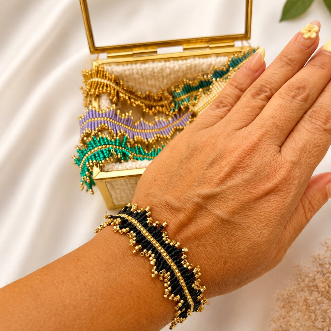 A close-up of a hand wearing a black and gold beaded wrap bracelet, with a matching decorative clutch bag and mirror in the background on a white satin surface.