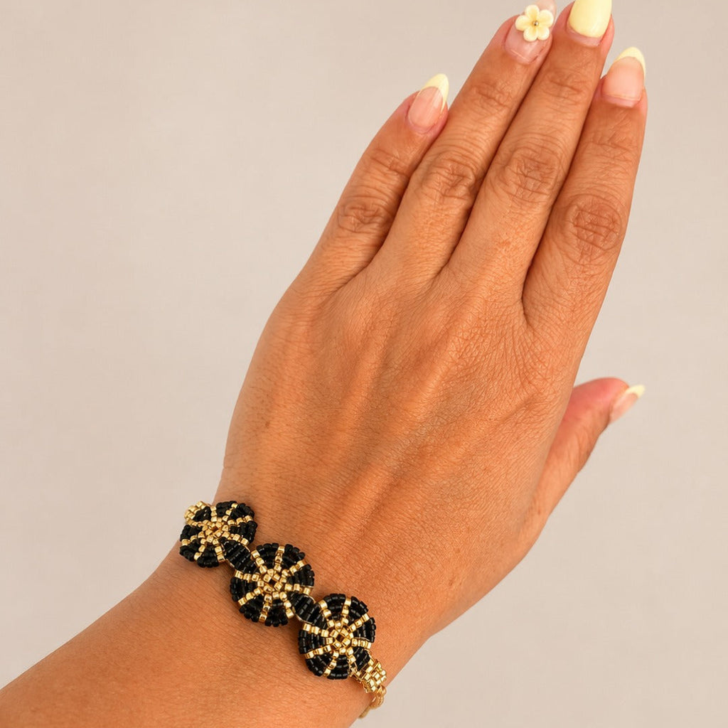 A close-up of a hand wearing a black beaded bracelet with gold starburst details, set against a plain light gray background.