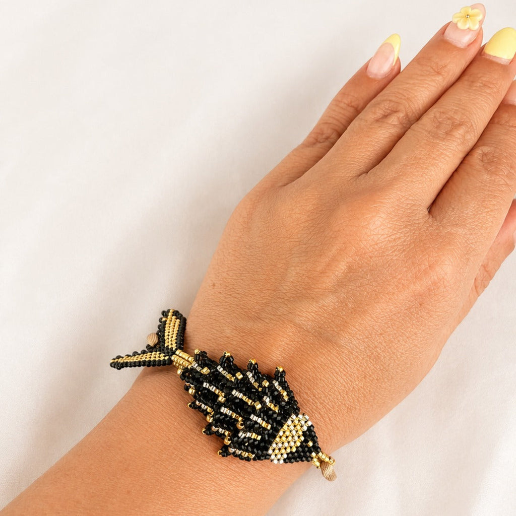 A hand and wrist wearing a beaded fish-shaped bracelet in black and gold seed beads against a plain white background.