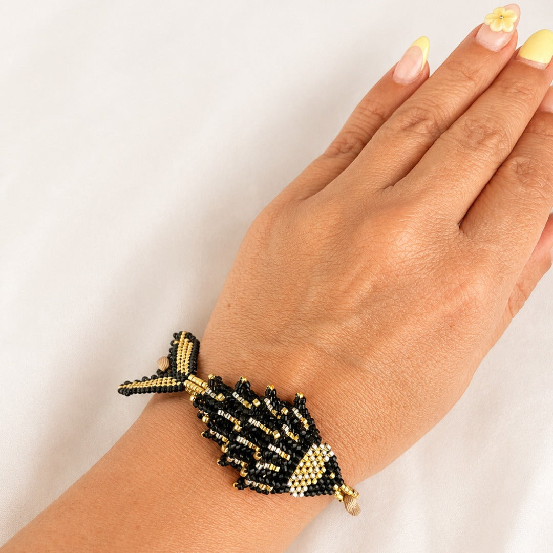 A hand and wrist wearing a beaded fish-shaped bracelet in black and gold seed beads against a plain white background.