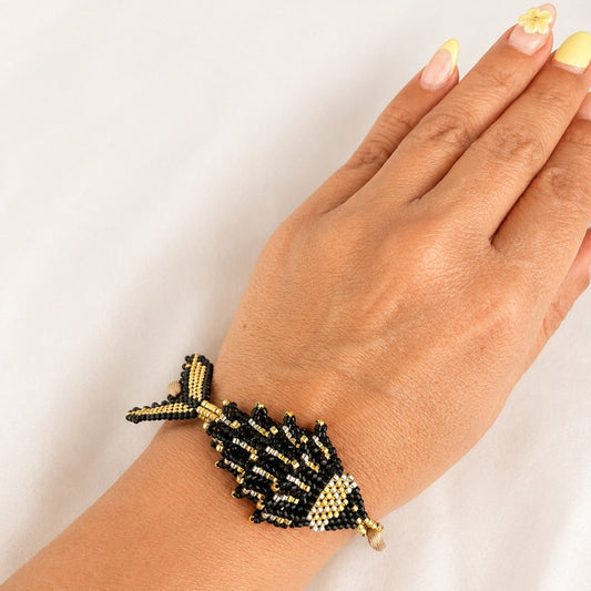A hand and wrist wearing a beaded fish-shaped bracelet in black and gold seed beads against a plain white background.