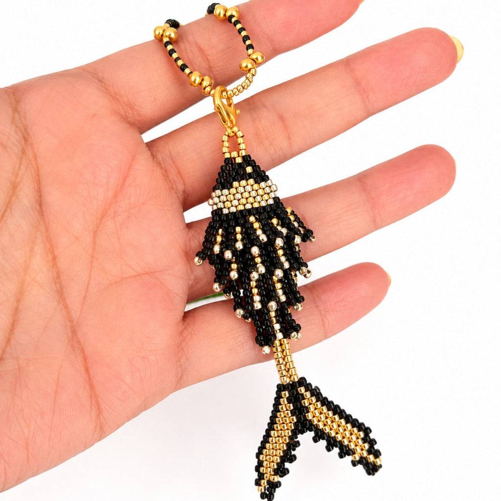 A hand holding a beaded fish-shaped keychain or bag charm in black and gold seed beads against a plain white background.