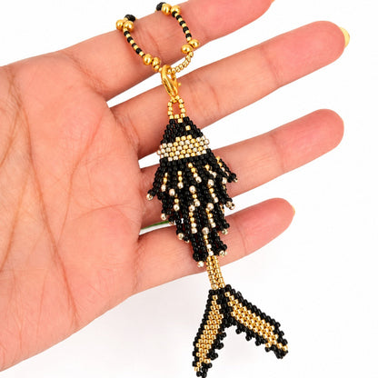 A hand holding a beaded fish-shaped keychain or bag charm in black and gold seed beads against a plain white background.