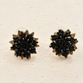 A pair of round stud earrings with a starburst design, featuring a black faceted crystal center surrounded by textured gold and bronze-toned petals, displayed on a light beige textured surface.