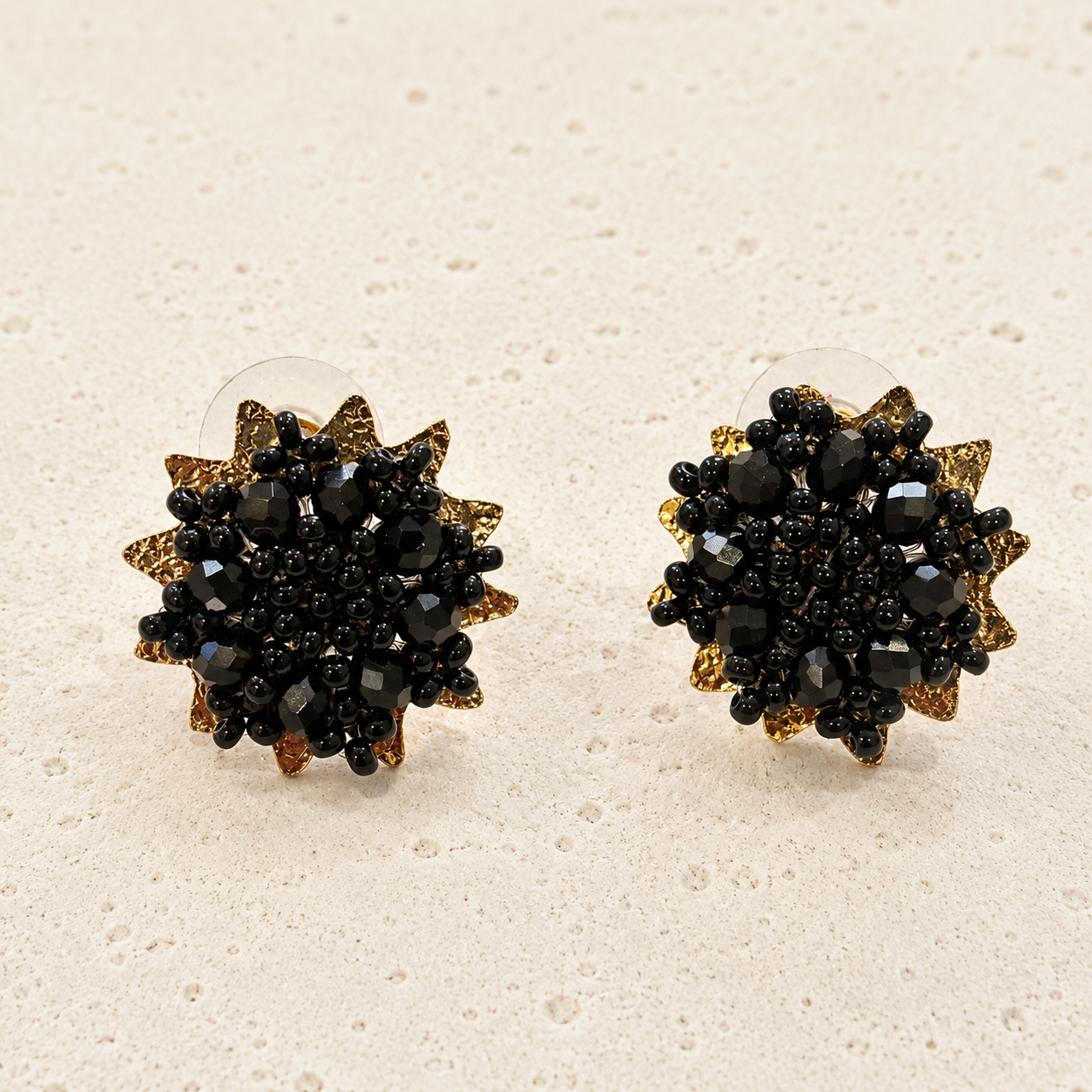A pair of round stud earrings with a starburst design, featuring a black faceted crystal center surrounded by textured gold and bronze-toned petals, displayed on a light beige textured surface.