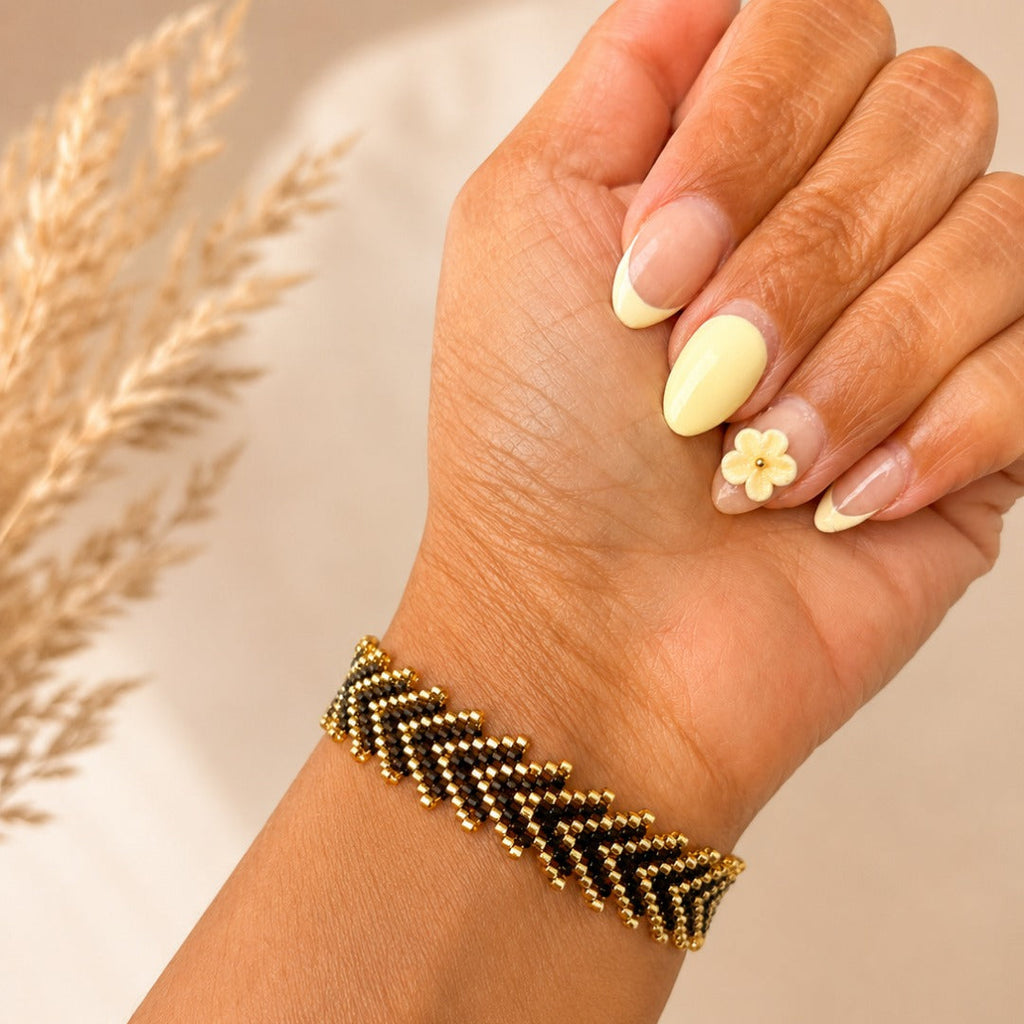 A close-up of a hand wearing a beaded chevron bracelet in black and gold, with neutral-toned manicured nails and a small flower accent, against a soft beige background with dried grass decor.