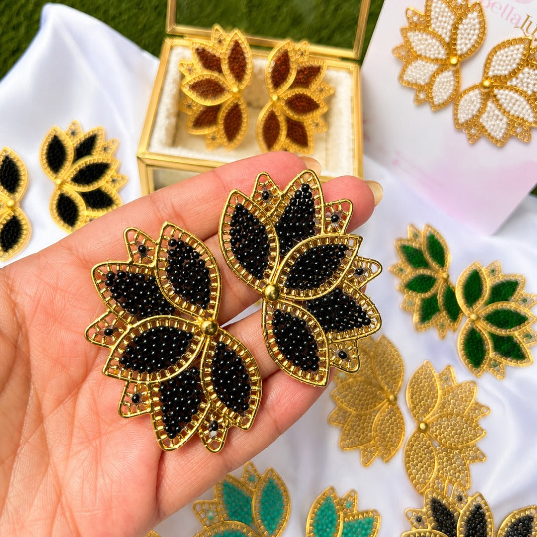 A hand holds a pair of gold-tone leaf-shaped earrings with black beadwork, with more matching earrings and a small display box arranged on white fabric in the background.