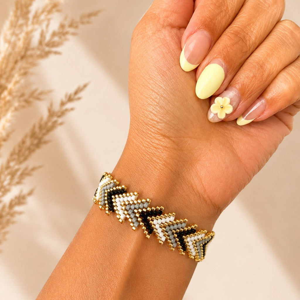 A close-up of a hand and wrist wearing a chevron-patterned beaded bracelet with gold, black, and white beads, set against a warm beige background with dried grass decor.