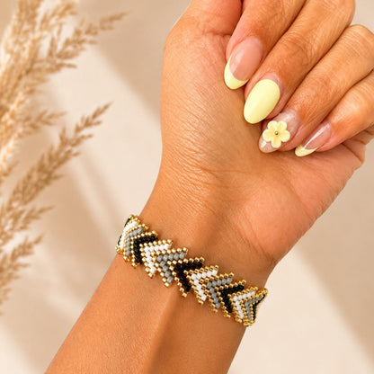 A close-up of a hand and wrist wearing a chevron-patterned beaded bracelet with gold, black, and white beads, set against a warm beige background with dried grass decor.