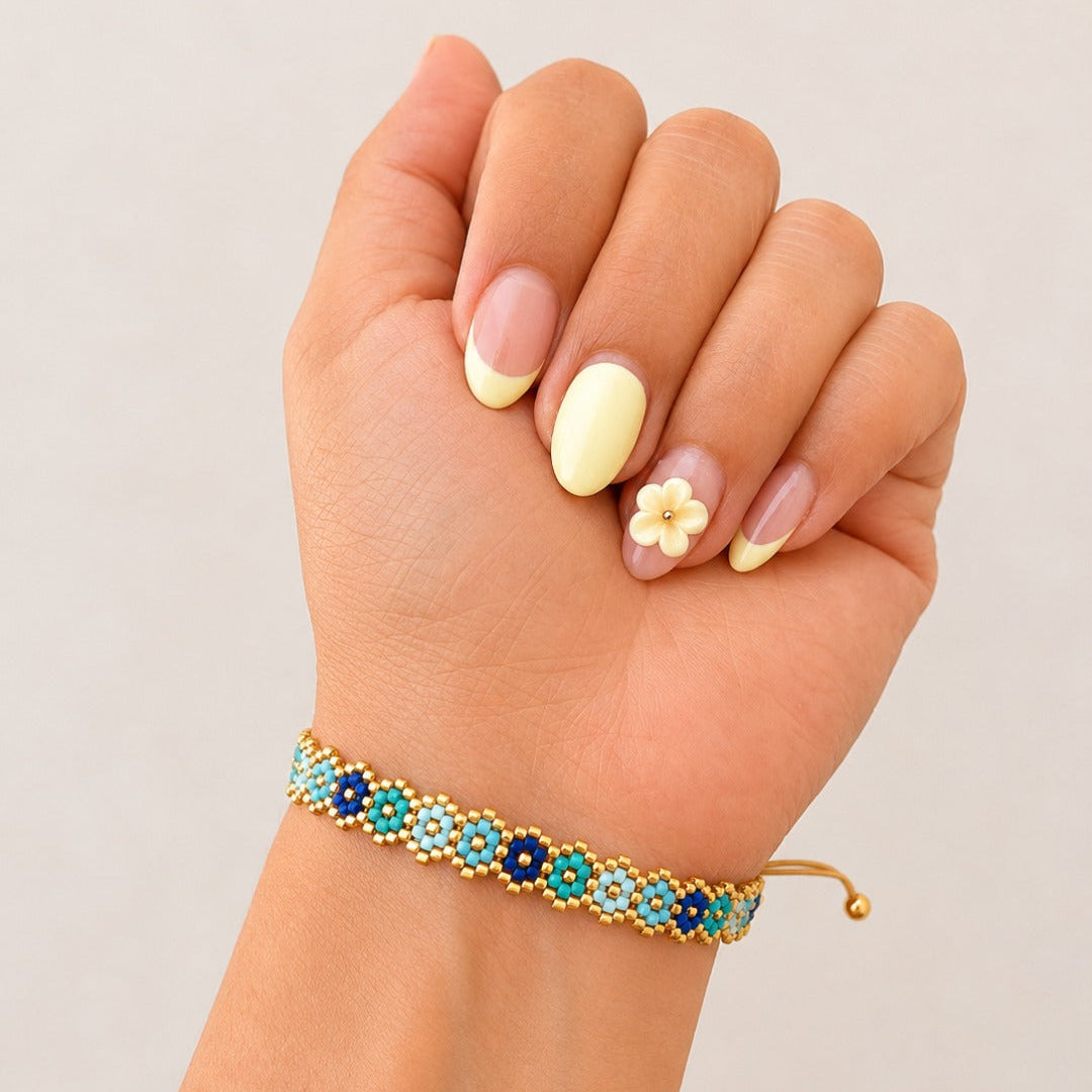 A close-up of a hand wearing a delicate gold bracelet adorned with blue and turquoise beads and small white flower details, set against a plain light gray background.