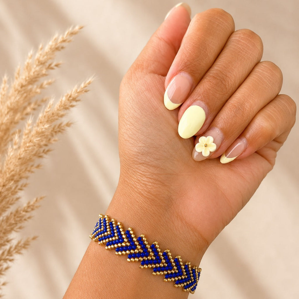 A close-up of a hand wearing a blue and gold chevron beaded bracelet, with neutral manicured nails and a small cream flower accent on one nail, against a soft beige background with dried grass decor.