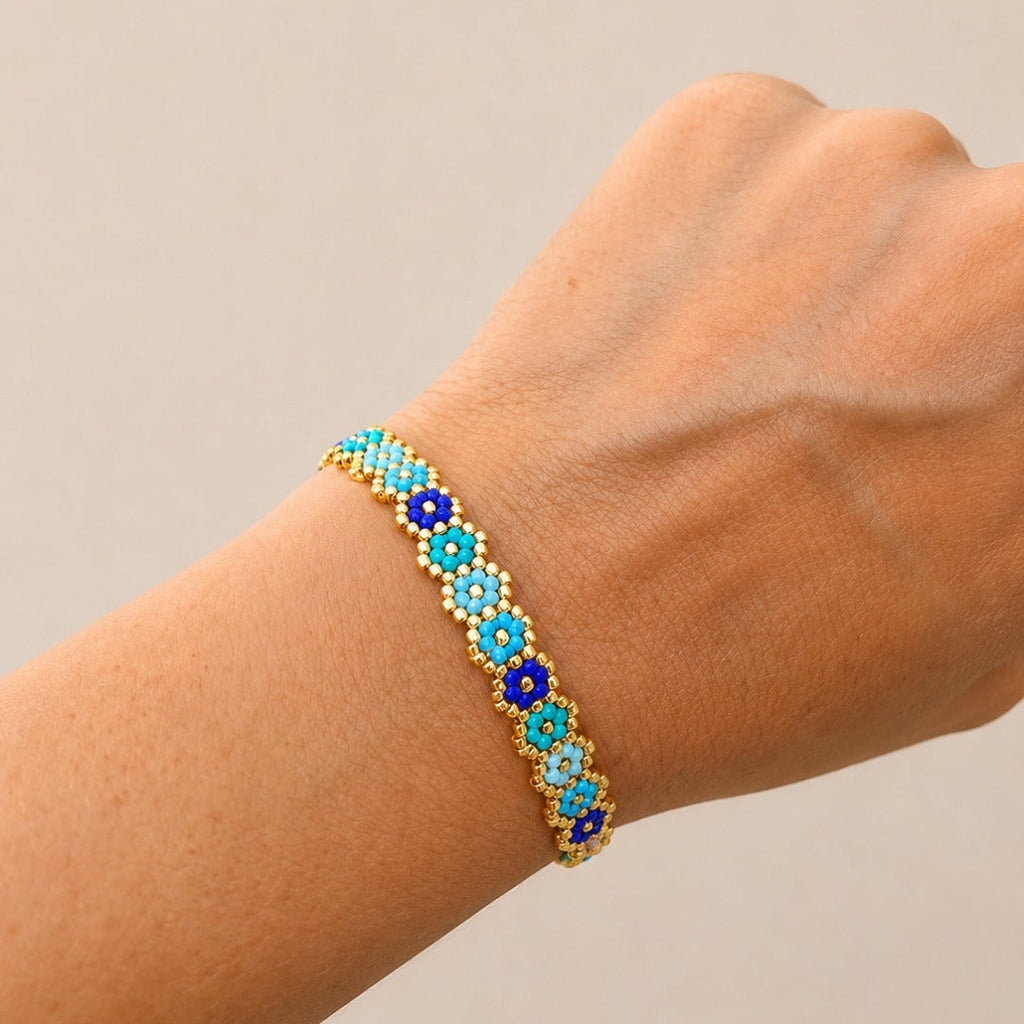 A close-up of a hand wearing a delicate beaded bracelet. The bracelet features repeating floral motifs with light blue and royal blue beads, accented by gold-tone metal borders and small white beads.