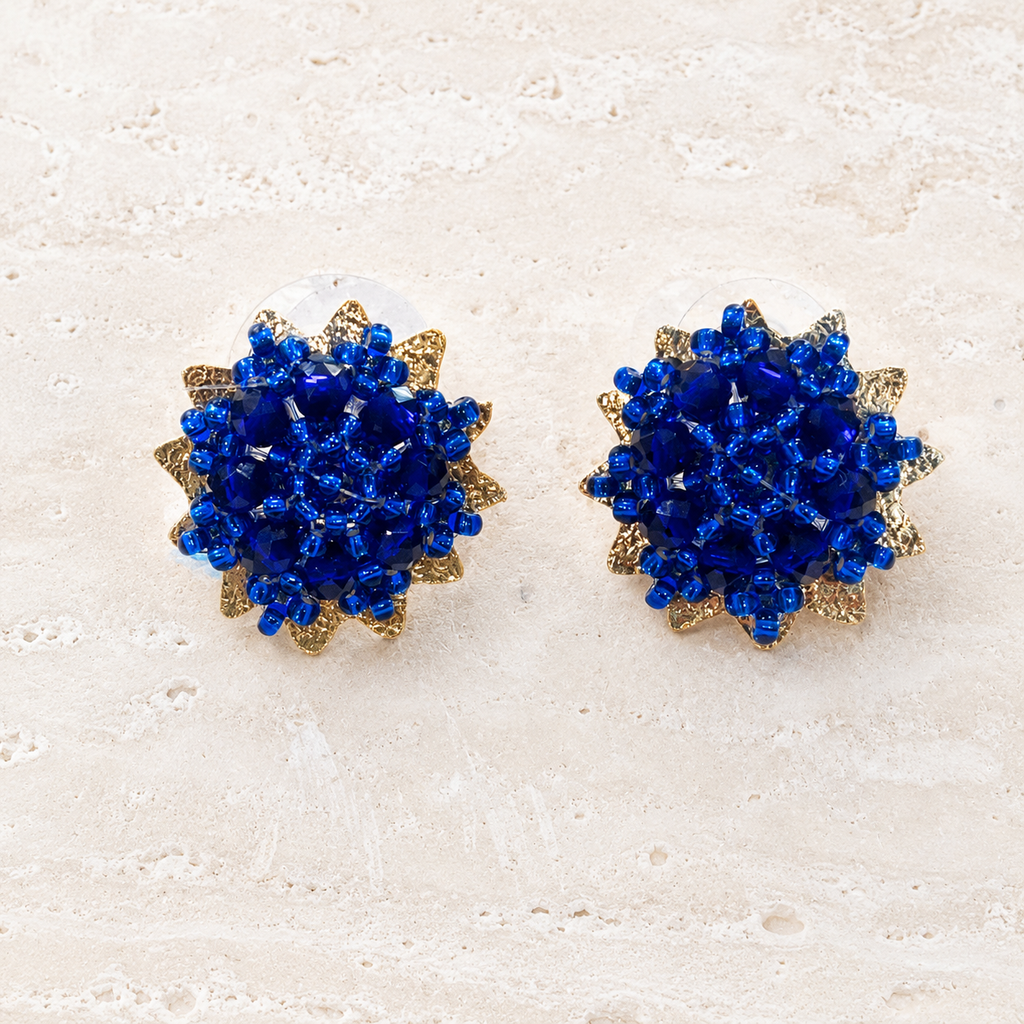 A pair of round stud earrings with deep blue faceted crystal clusters surrounded by textured gold-tone metal petals, displayed on a light beige stone surface.