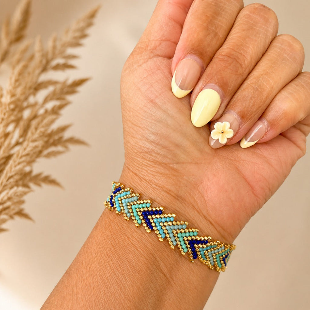 A close-up of a hand wearing a beaded chevron bracelet with navy blue, turquoise, and gold beads, set against a warm beige background with dried grass.
