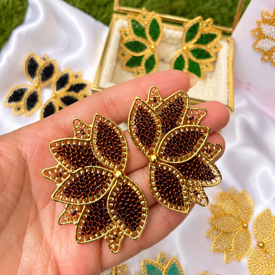 A hand holds a pair of gold-tone leaf-shaped brooches decorated with dark brown seed beads and small red accents, with more matching jewelry and an open display box in the background on white satin fabric and green grass.