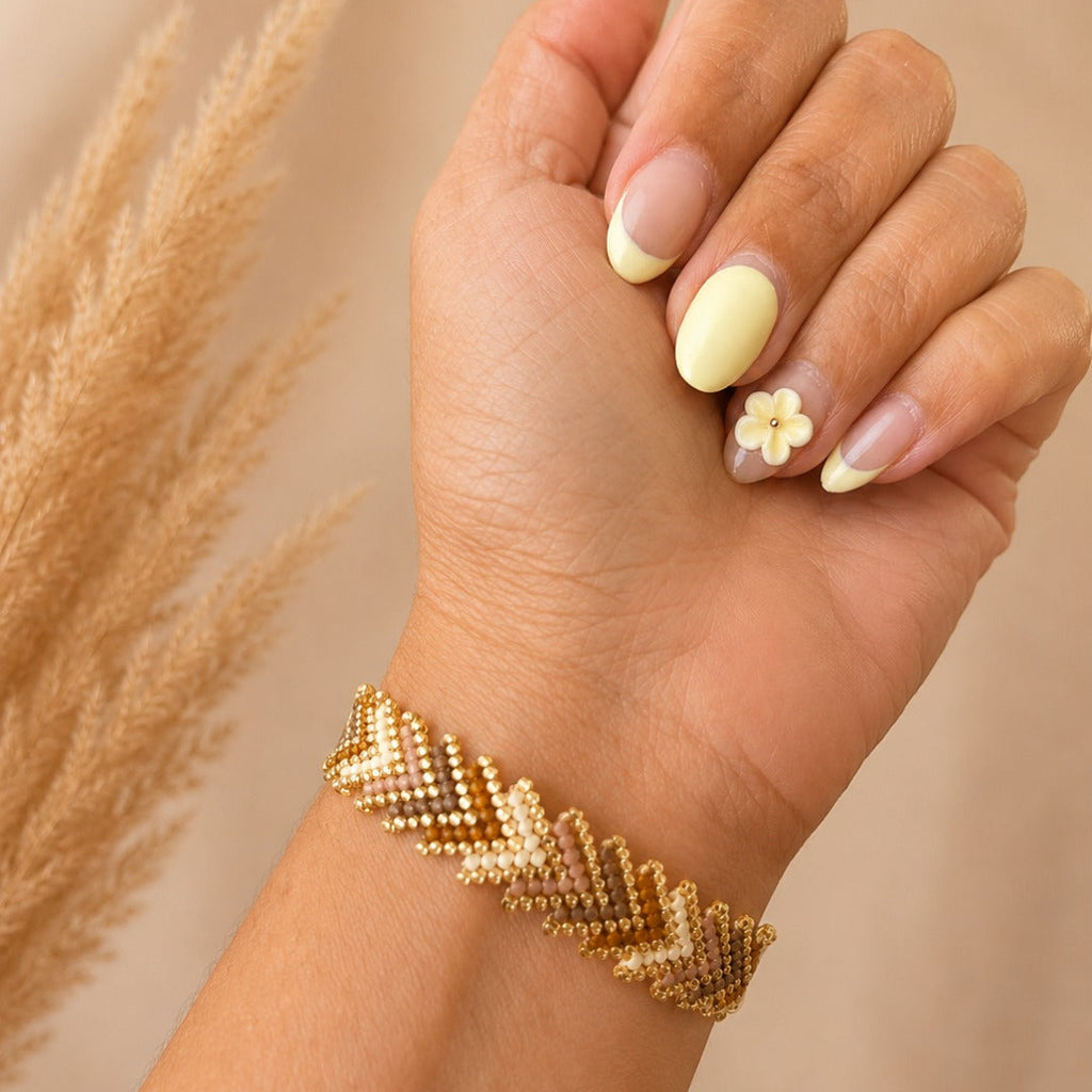 A close-up of a hand wearing a gold chevron-pattern beaded bracelet, with French-tipped nails and a small flower nail art accent, against a soft beige background with dried grass.