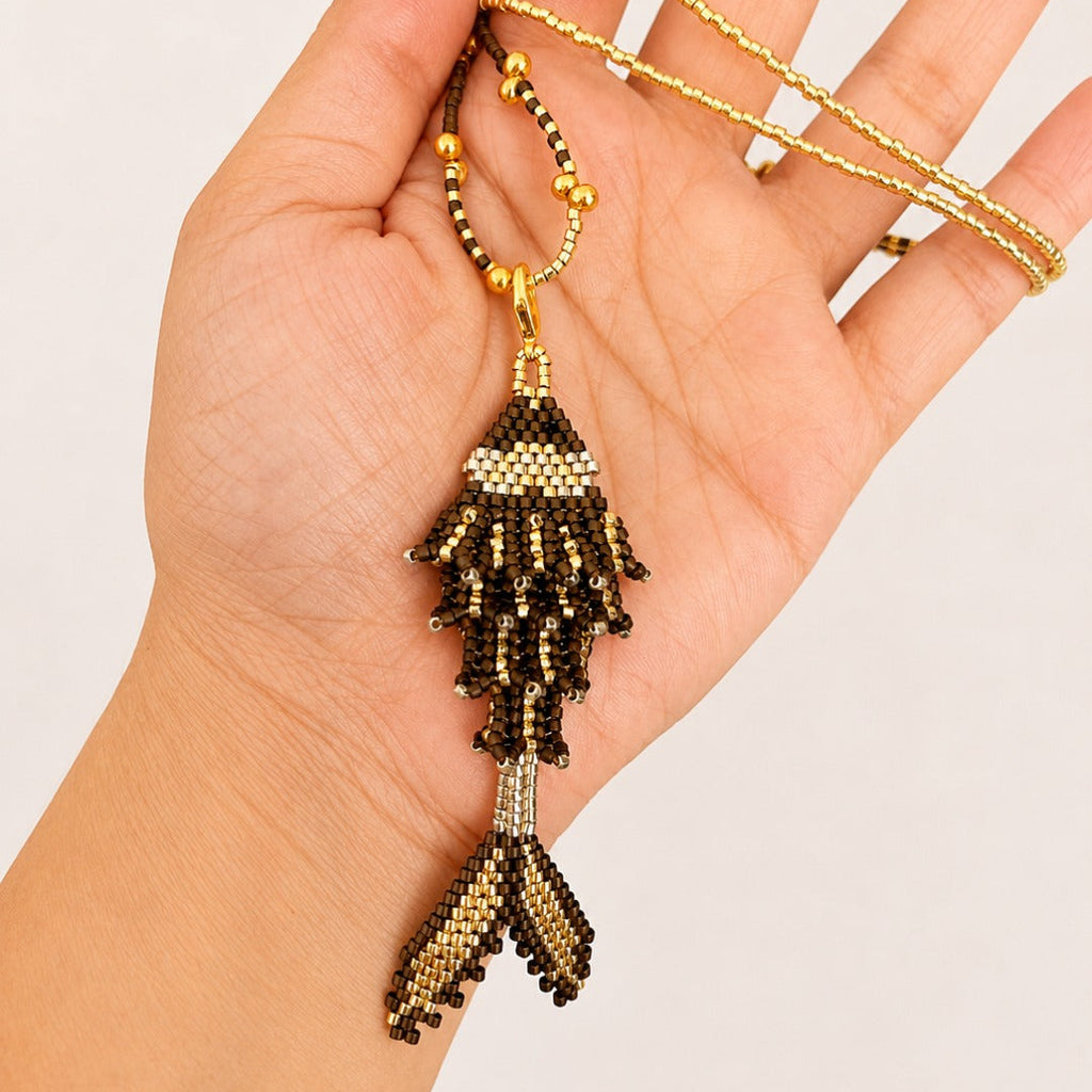 A hand holding a gold and dark brown beaded fish pendant necklace against a plain light background.