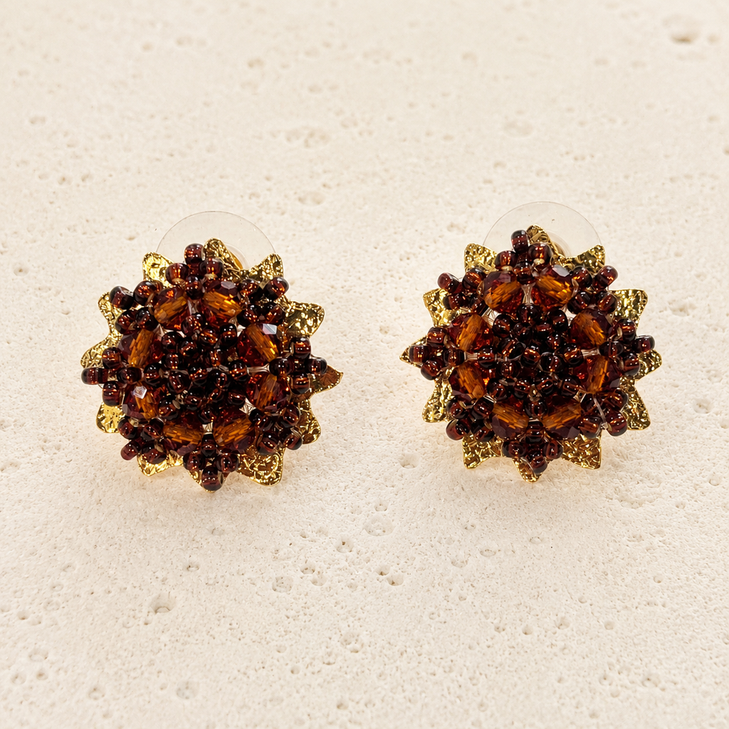 A pair of round cluster stud earrings featuring faceted amber-brown crystals arranged in a floral or sunburst pattern with textured gold-toned edges, shown on a light beige textured surface.