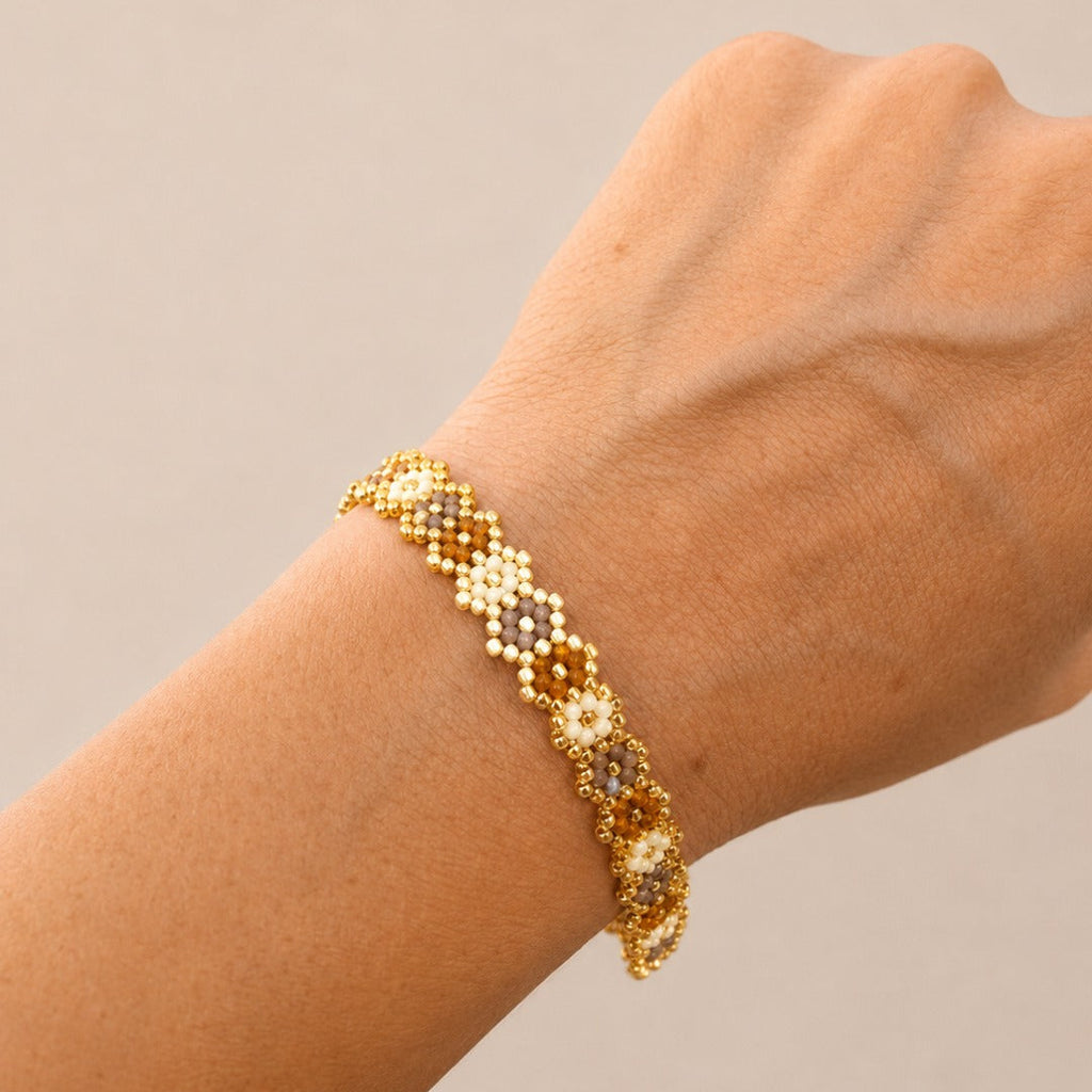 A gold beaded bracelet with floral details, shown worn on a person's wrist against a neutral beige background.