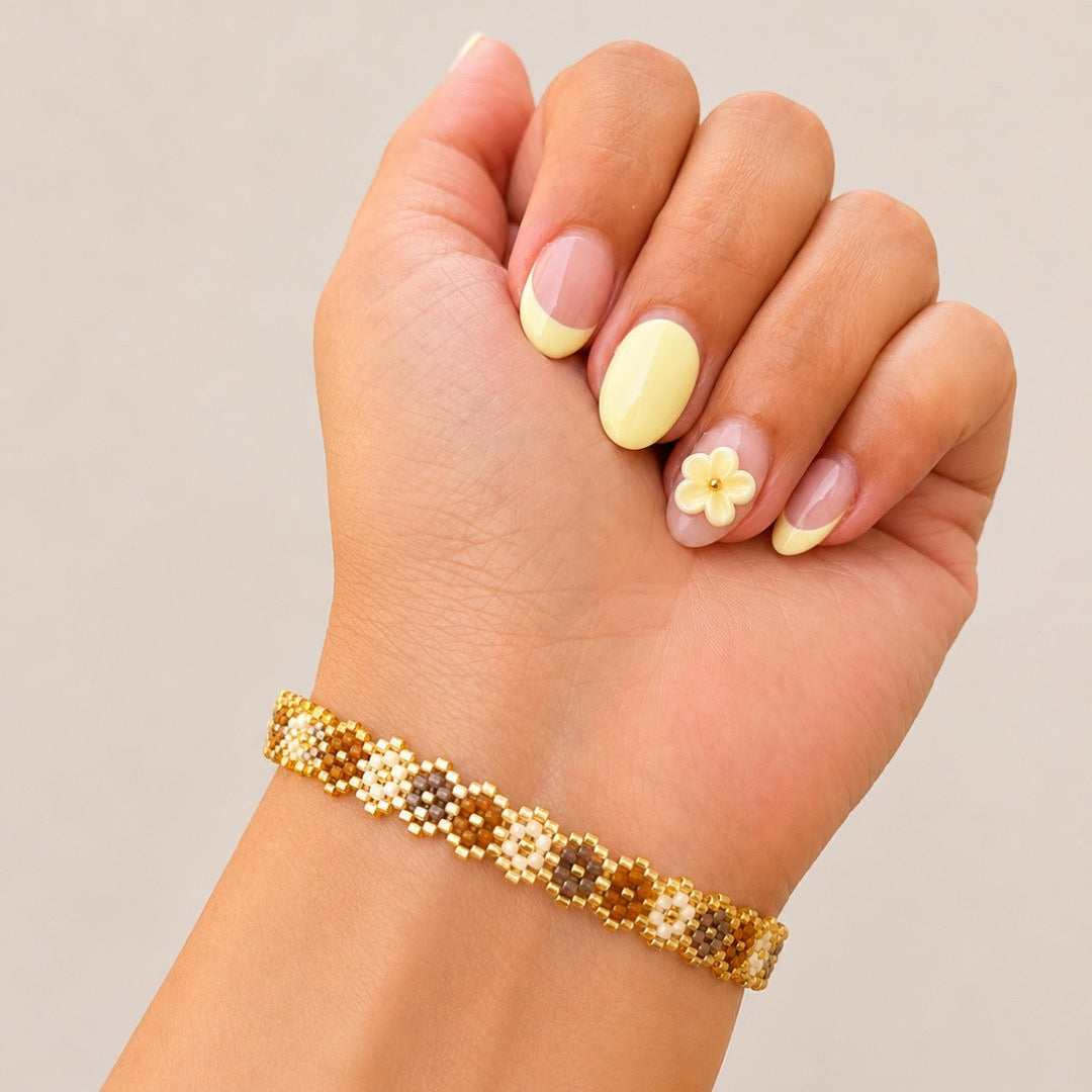 A close-up of a hand wearing a delicate beaded bracelet. The bracelet features a gold-tone chain with woven segments of brown and gold seed beads.